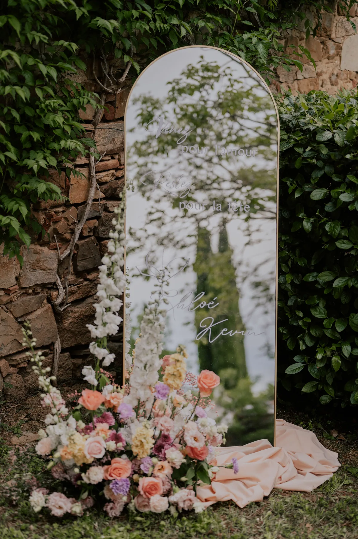 Grand miroir arche gravé, entouré de fleurs, installé contre un mur végétal