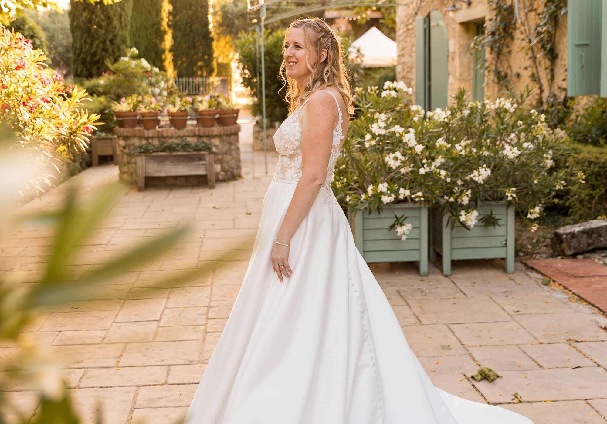 La mariée se retourne, traîne déployée, dans le patio fleuri du domaine