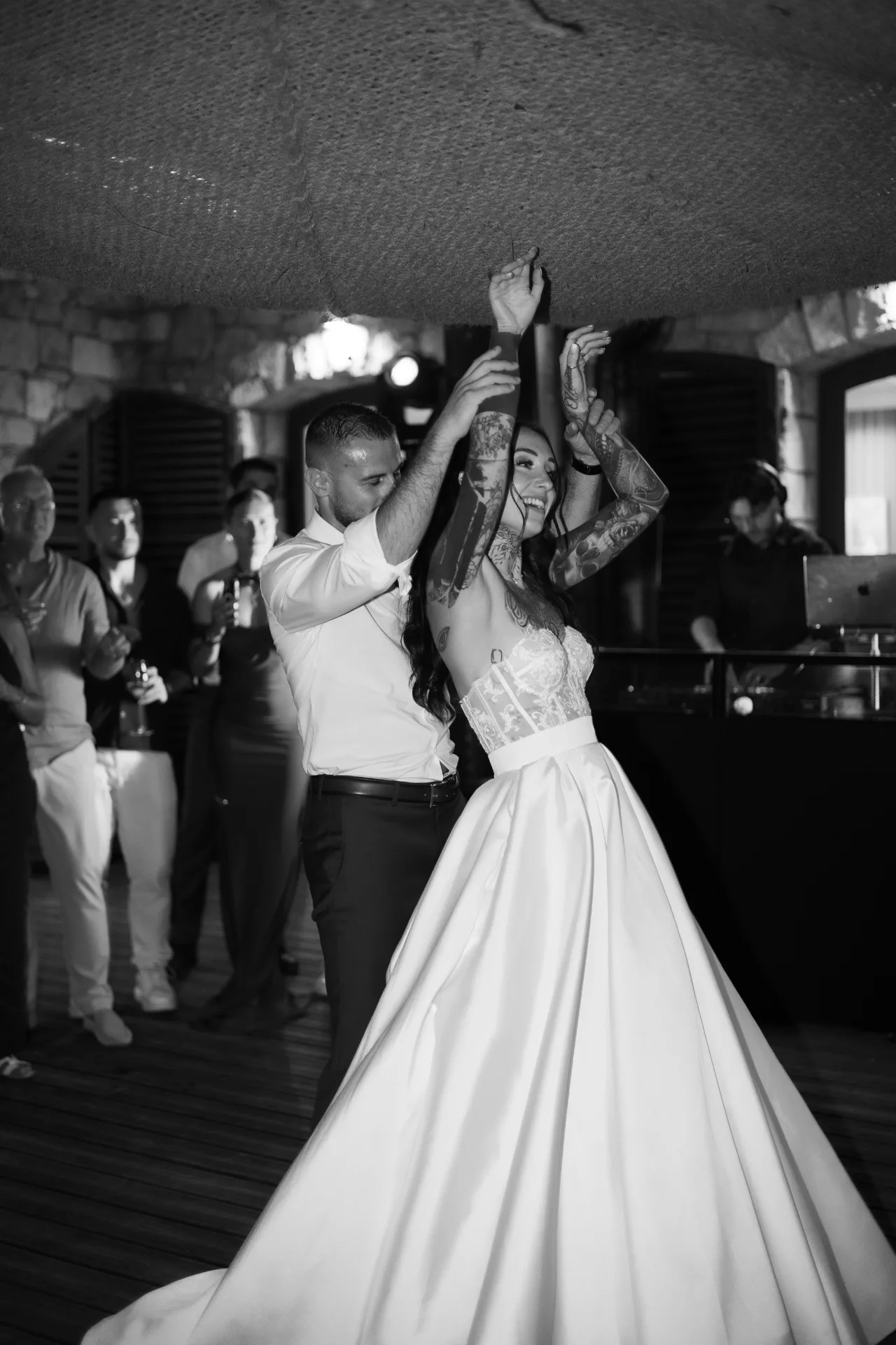 Les mariés dansent, bras levés, entourés de leurs invités, en noir et blanc